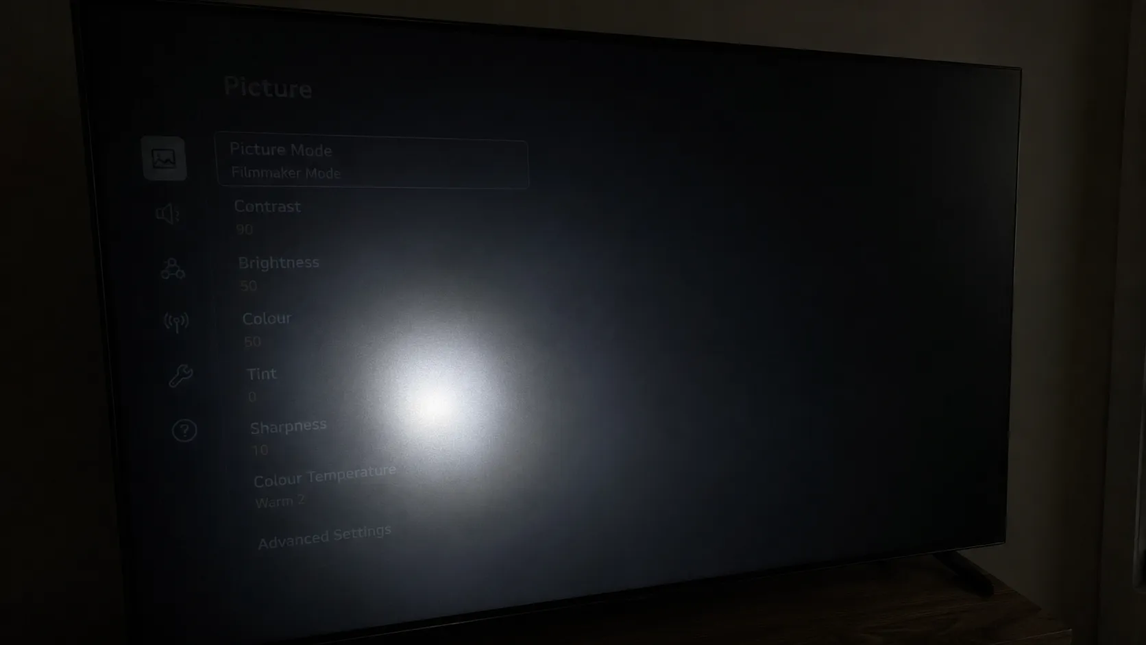 Original photo of a flashlight test on a 55-inch TV screen in a dark room, showing a faint settings menu visible at an angle with flashlight glare highlighting screen uniformity.