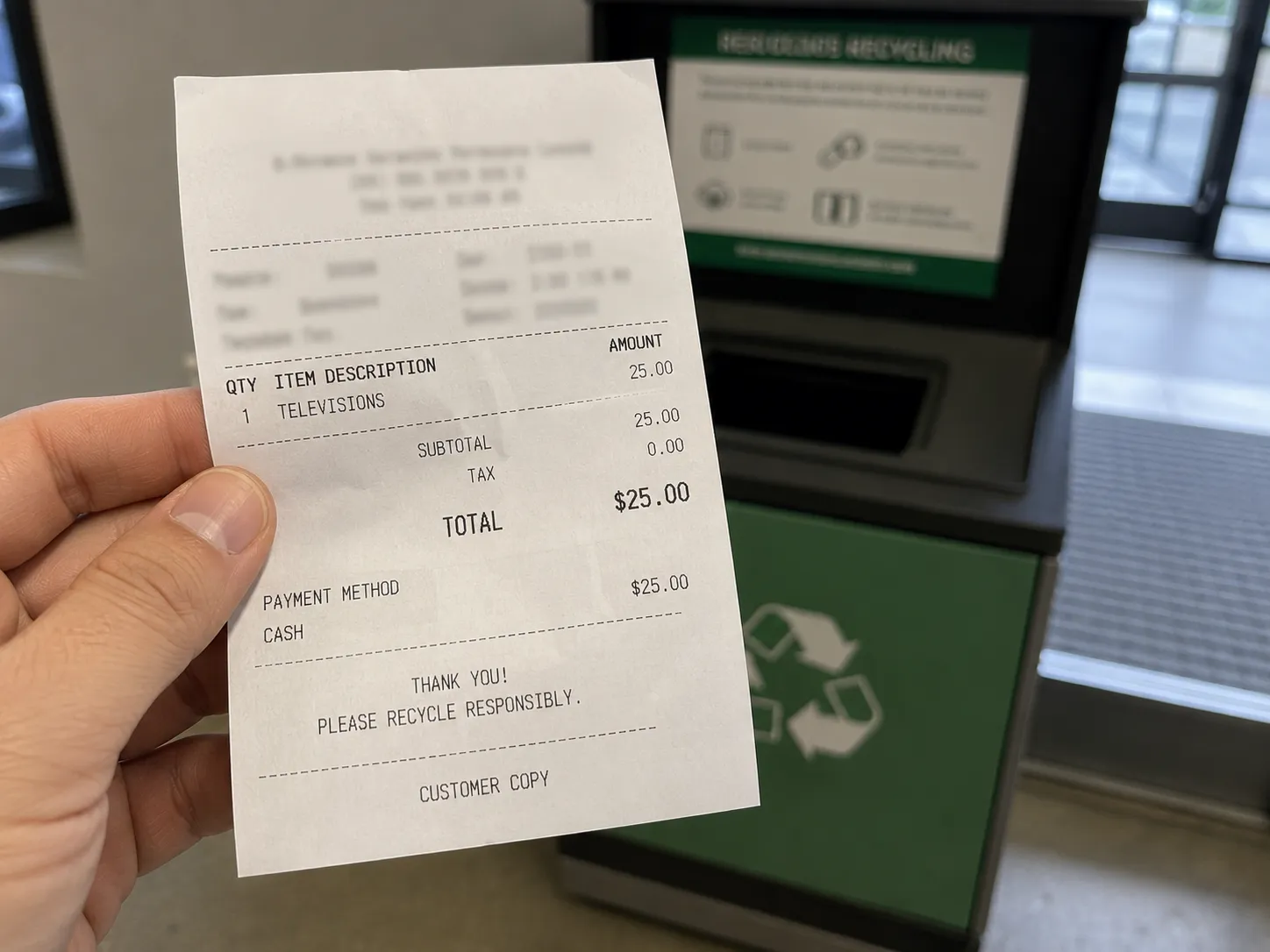 Hand holding an electronics recycling drop-off receipt near a TV recycling collection bin, showing the recycling fee for disposing of a broken flat-screen television.