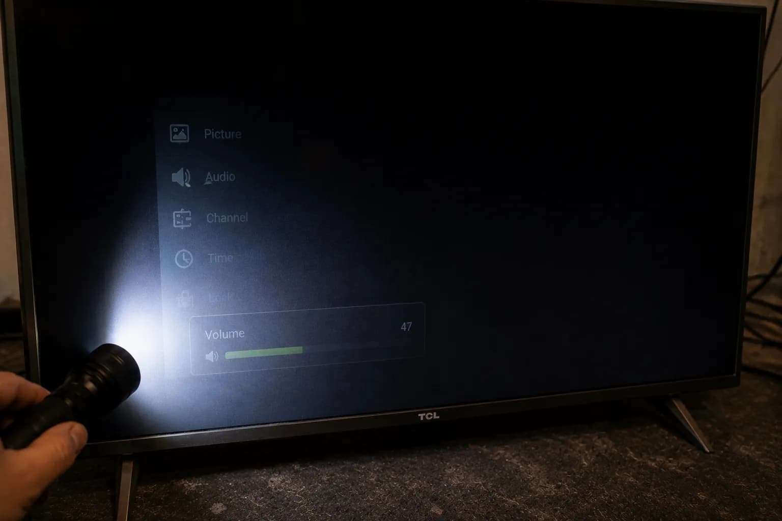 Flashlight reveals a faint menu and active volume bar on a dark TCL TV screen, confirming the LCD panel works while the LED backlight has failed.