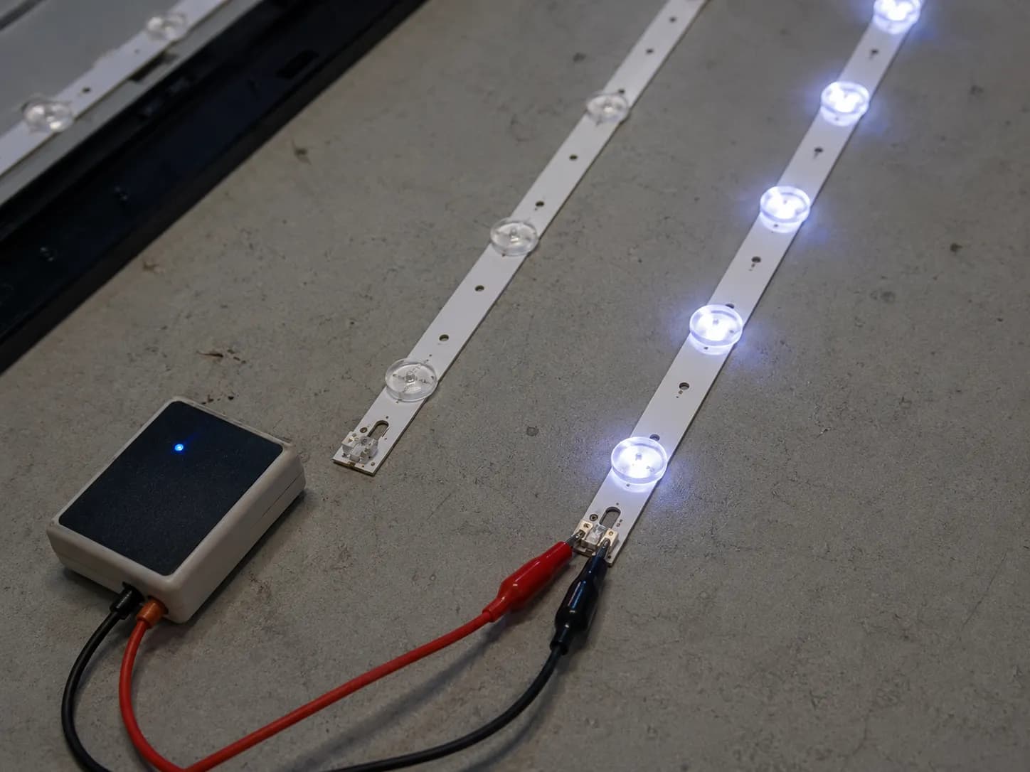LED backlight tester connected to a Samsung TV LED strip with one strip glowing bright and another remaining dark, demonstrating how to identify a faulty TV backlight strip.