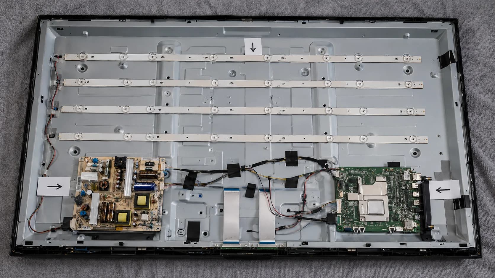 Overhead view of an opened flat-screen TV showing LED backlight strips, power board, main board, and ribbon cables with arrows indicating the main internal components.