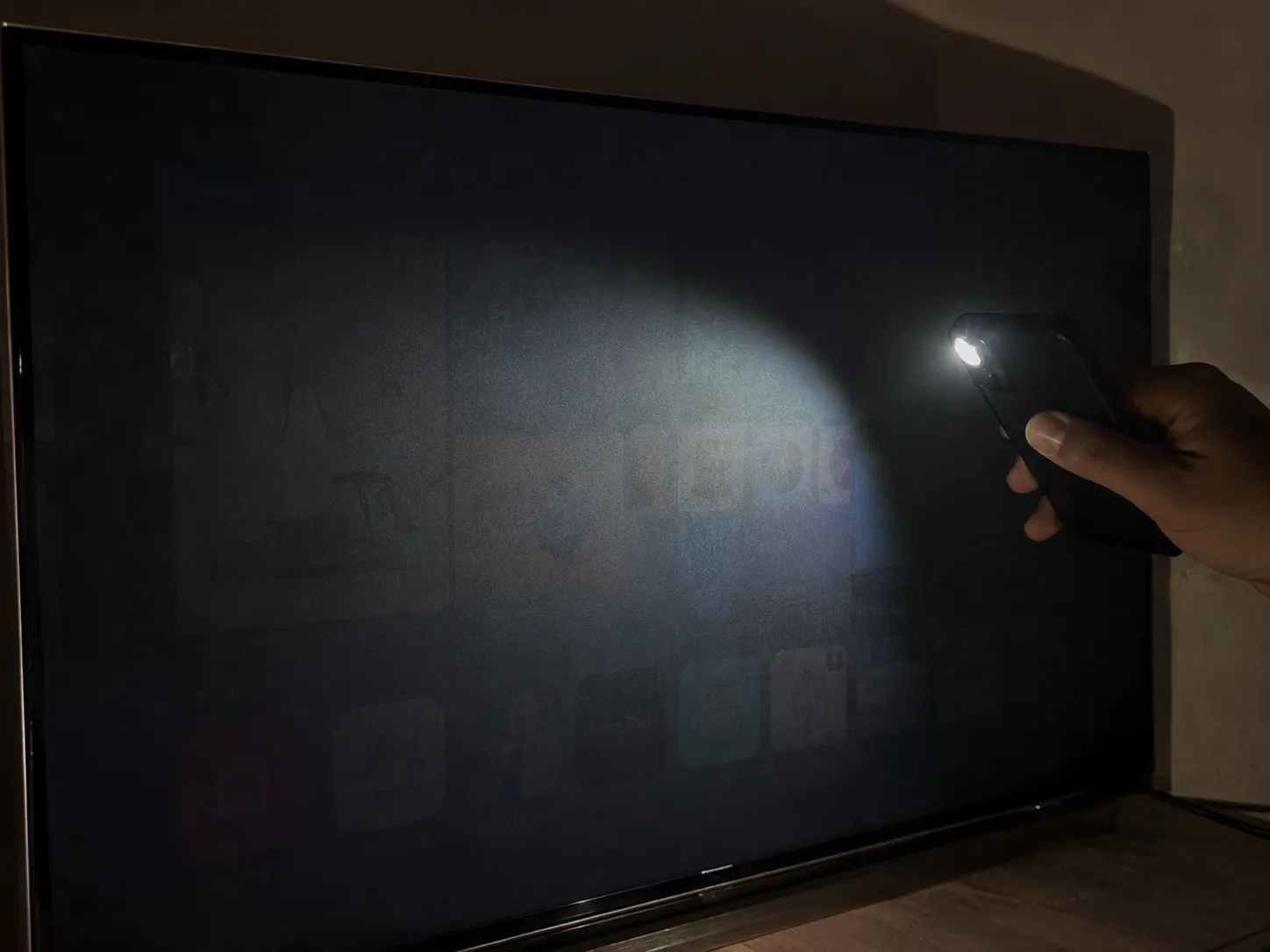 Phone flashlight shining on a dark Samsung TV screen revealing a faint home screen image, demonstrating a failed LED backlight during the flashlight diagnostic test.