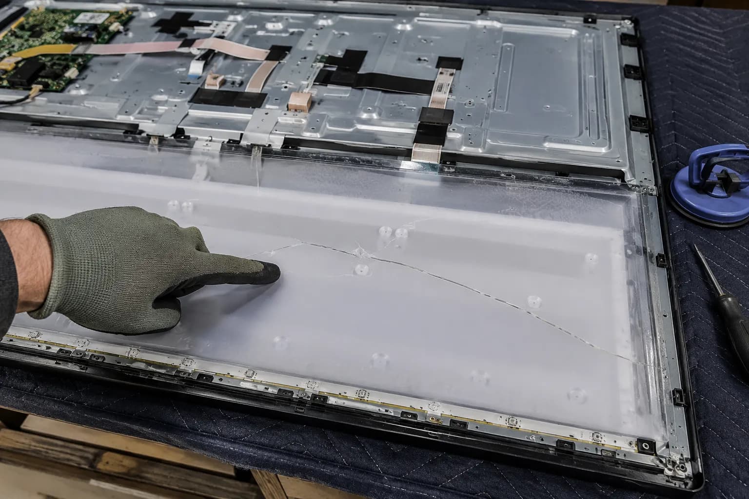 Opened flat-screen TV with the rear cover removed, showing a crack beneath the diffuser layer as a gloved hand points to the damaged panel during screen replacement inspection.