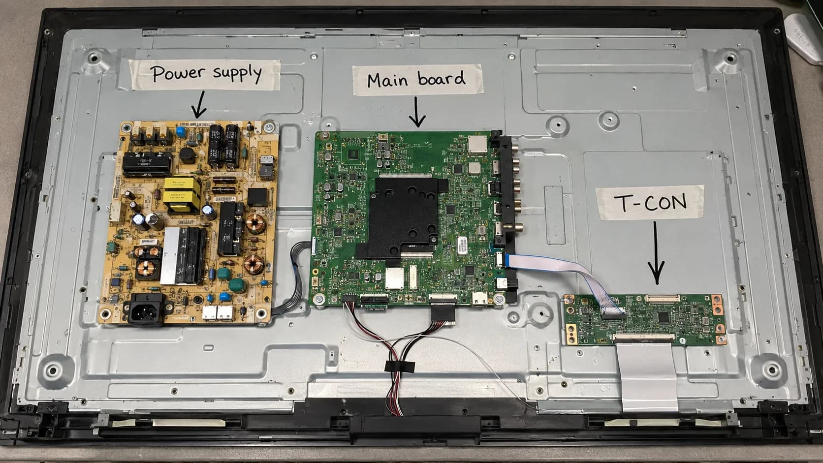 Annotated teardown of an open 55-inch TCL TV showing the power supply, main board, and T-CON board with labeled arrows and ribbon cable connections inside the rear chassis.