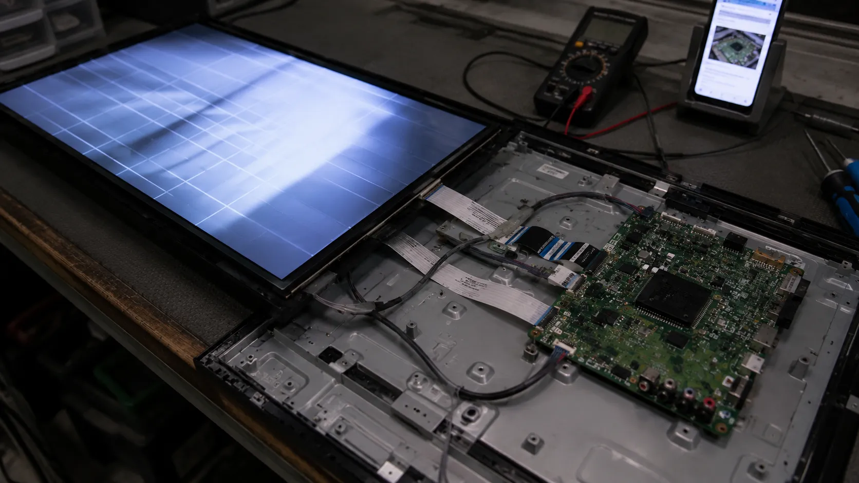 Open TV powered on face-down during a bench test with a lit test pattern, exposed main board, multimeter, and phone used to verify ribbon cable connections before reassembly.