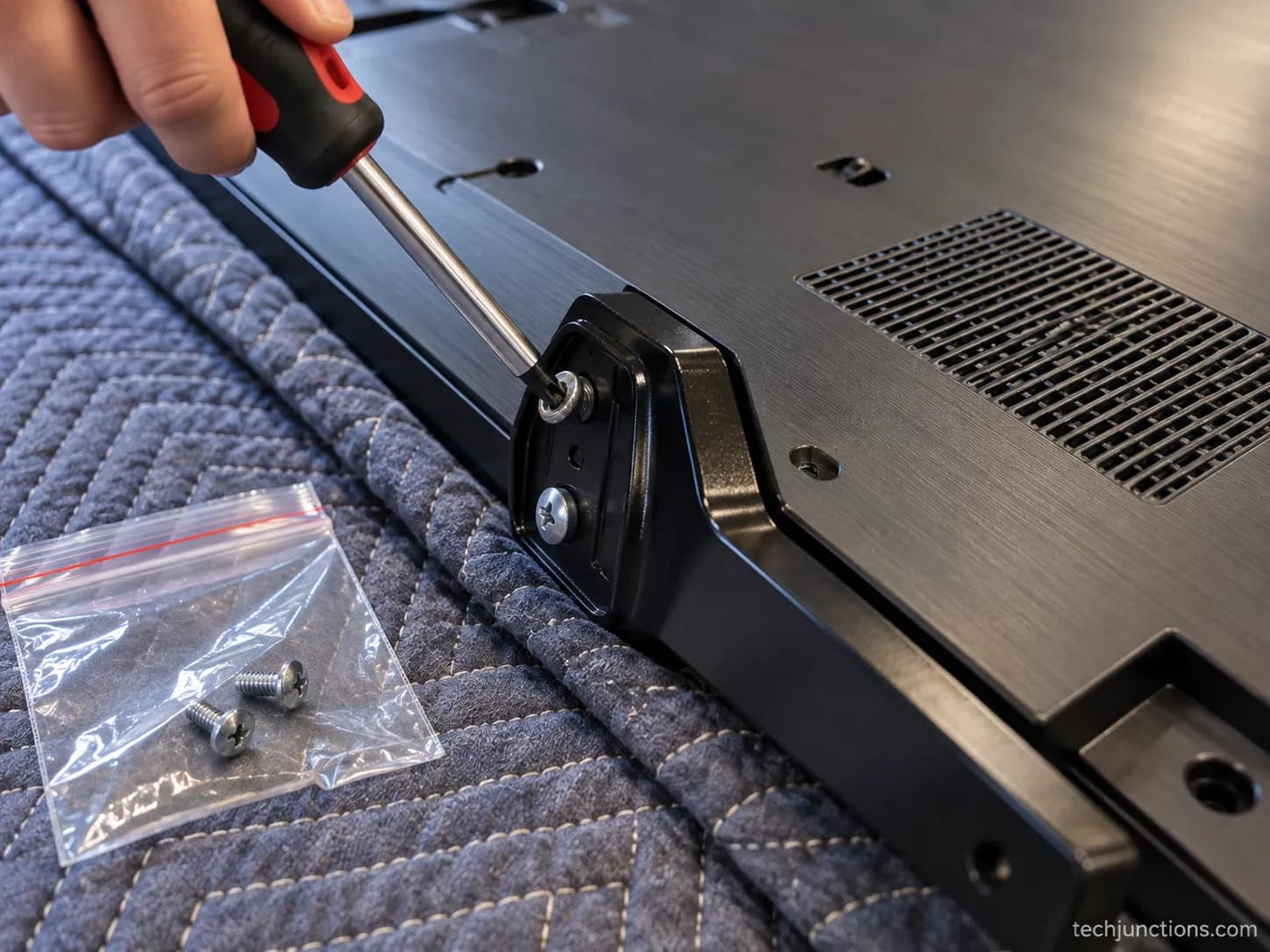 Hand using a PH2 Phillips screwdriver to remove a screw-attached LG TV stand leg, with removed screws bagged beside it.